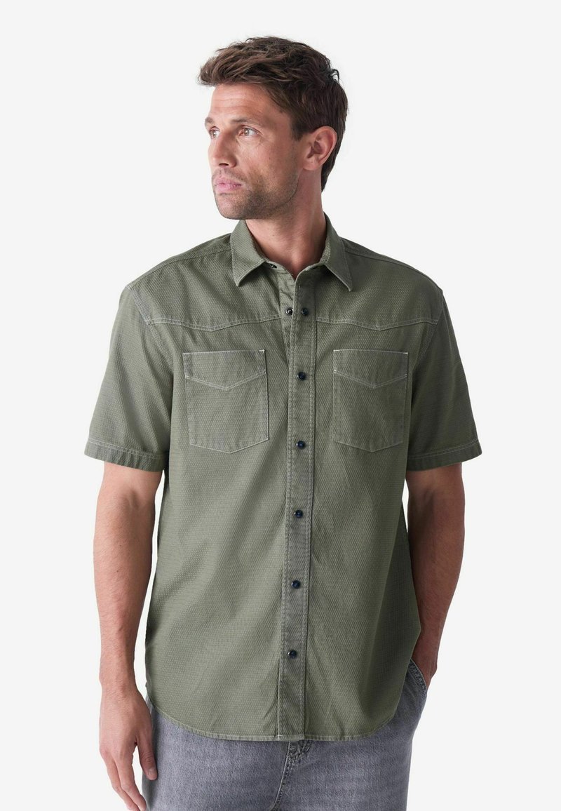 Camisa de manga corta en verde oliva, con botones y un patrón texturizado, dos bolsillos en el pecho y botones oscuros. Dobladillo ligeramente curvado y ajuste casual.