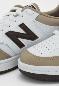 Vita och beige sportskor har en perforerad tåhuvud, bruna detaljer och en framträdande brun "N"-logotyp på sidan. Snörena är vita.