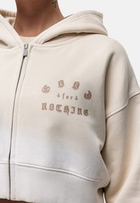Hoodie cropped beige avec une fermeture éclair à l'avant, arborant un imprimé graphique marron foncé portant la mention "GOOD for NOTHING." Tissu doux, coupe décontractée.