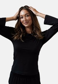 Czarny długi rękaw top z miękkiego materiału, z okrągłym dekoltem i luźnym krojem. Noszony z rękami opartymi na głowie, ukazujący naturalne włosy.