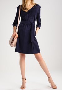 Námořnická modrá zavinovací šaty s výstřihem do V, tříčtvrtečními rukávy a látkovým opaskem. Vytvořeno s lehkým růžovým crossbody batohem a béžovými sandály na podpatku.