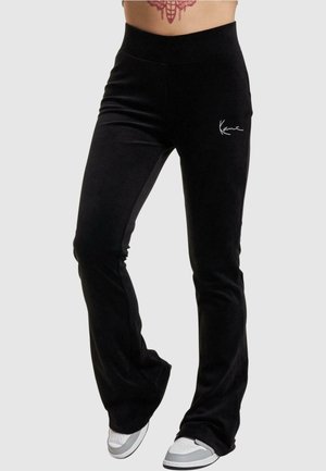 Persona que lleva pantalones negros acampanados con un logo blanco en el muslo y zapatillas blancas, mostrando un tatuaje en el abdomen contra un fondo liso.