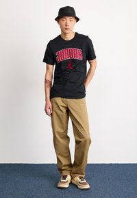 Černé tričko Jordan s červeným nápisem, béžové kalhoty a vícebarevné tenisky. Model nosí černý klobouk baskytář. Jednoduchý, ležérní outfit.