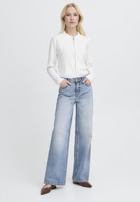 Femme blonde debout, les mains dans les poches, portant un pull à fermeture éclair blanc, un jean large bleu clair et des ballerines pointues marron.