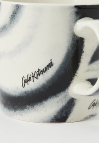 Taza de cerámica con un patrón de remolinos en blanco y negro, con un asa redondeada y el texto "Café Kitsuné" en letra cursiva negra en el lateral.