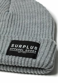 Siva rebrasta pletena kapa s crnom etiketom na kojoj bijelim slovima piše "SURPLUS APPAREL GOODS EST. 1997." Mekana tekstura.