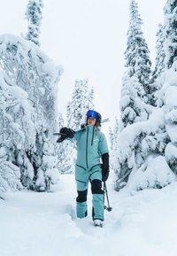 Kombinezon narciarski w kolorze morskim z czarnymi akcentami, kieszeniami na suwak i rękawiczkami. Narciarz trzymający kijki narciarskie w zaśnieżonym lesie z wysokimi drzewami iglastymi.
