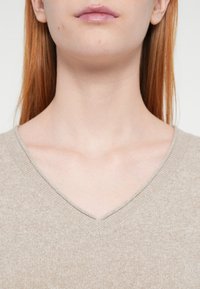 Pull en V beige clair, avec une texture douce et un motif tricoté. L'encolure souligne subtilement la zone de la clavicule.