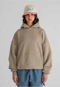 Beige overdimensjonert hettegenser med kengurulomme og ribbede mansjetter, sammen med blå shorts med en rosa brodert logo. Kappen har pastellfarger.