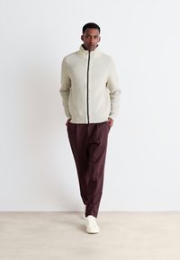 Homme marchant les mains dans les poches, portant un pull zippé crème, un pantalon marron foncé et des baskets blanches sur un fond blanc uni.