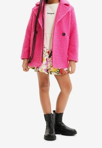 Roze gestructureerde jas van nepbont met reverskraag, twee zwarte knopen, gedragen over een bloemenjurk. Zwarte enkellaarsjes met ritssluitingen aan de zijkant.