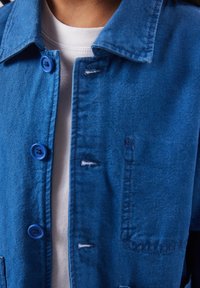 Gros plan d'une personne portant une chemise bleue boutonnée sur un T-shirt blanc, montrant le col, les boutons et une partie de la poitrine.