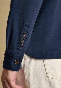 Marineblaues Langarmhemd mit Knöpfen an den Manschetten, versehen mit goldener "JOULES"-Stickerei. Der Stoff ist glatt und das Design ist figurbetont.