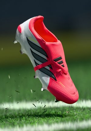 Helles pinkfarbenes Fußballschuh mit einer strukturierten Oberseite, schwarzen Streifen an der Seite und einer weißen Sohle. Stollen sind unter dem Schuh sichtbar.