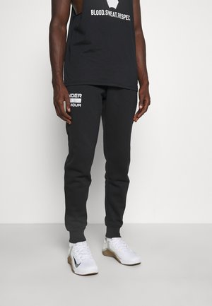 Pantaloni da jogging neri con il testo "UNDER ARMOUR" in bianco sulla gamba sinistra, abbinati a una maglietta senza maniche nera e sneakers bianche.