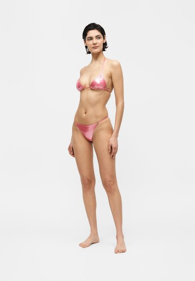 Nő áll mezítláb, csillogó rózsaszín háromszög topból és alsóból álló bikinit viselve, enyhén oldalra néz egy egyszerű világos háttér előtt.