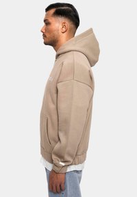 Beige hættebluse med en kengurulomme, ribbede manchetter og kant. Blødt stof, afslappet pasform. Modellen har lyseblå jeans og en hvid skjorte under.