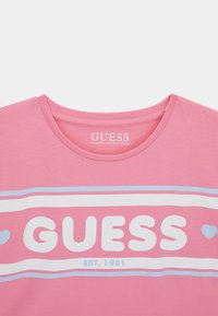 Roosa orgaanilisest puuvillast T-särk ümmarguse kaelusega, millel on "GUESS" logo valgete ja siniste triipudega ning allosas asuv asutamisaasta "1981".