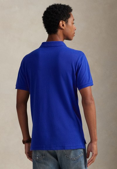 Háttal álló férfi, aki királykék rövid ujjú pólót és világoskék farmert visel, egyszínű bézs háttér előtt.