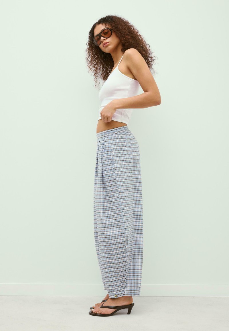 Femme aux cheveux bouclés portant des lunettes de soleil, un débardeur blanc, un pantalon large à carreaux ample et des sandales noires à talons, debout de profil.