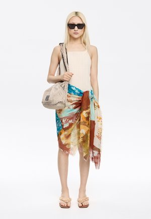Body beige côtelé, foulard fleuri multicolore noué à la taille, sac en filet beige, lunettes de soleil noires et tongs beiges avec des brides texturées.
