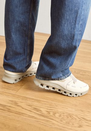 Weisse Sneakers mit strukturiertem Obermaterial, ausgestattet mit derben Sohlen und Ausschnitten. Getragen mit blauen Jeans und stehend auf einem Holzfußboden.