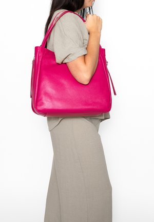 Donna con outfit beige che tiene una borsa a tracolla in pelle rosa acceso su sfondo bianco.