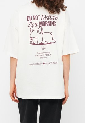 Personne portant une chemise blanche avec une illustration d'un chien endormi et un texte sur les matins lents et à ne pas déranger avant 11h.