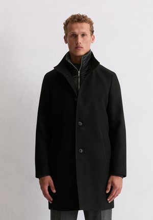 Mann trägt einen schwarzen Knopfmantel über einer schwarzen Reißverschlussjacke und steht vor einem einfarbigen hellen Hintergrund.