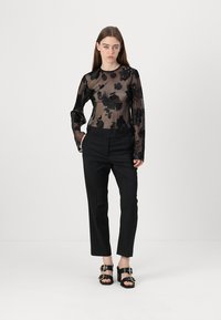 Svart gennemsigtig langærmet top med blomsterbroderi, parret med skræddersyede sorte bukser og kraftige sorte sandaler med gyldne hardware-detaljer.
