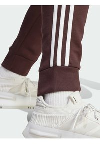 Pantaloni tuta color bordeaux con accenti bianchi a tre strisce sui polsini, abbinati a sneakers bianche a trama e calzini bianchi a coste.