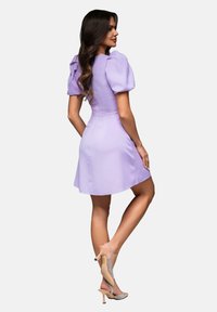 Robe lilas avec des manches bouffantes, un corsage ajusté et une jupe évasée. Portée avec des talons nus ornés de bijoux en strass. Texture de tissu lisse.