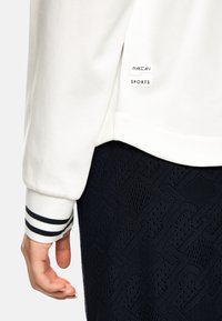 Weißes Sportoberteil mit Streifendetails an den Bündchen, versehen mit einem Etikett mit der Aufschrift "Marc Cain Sports". Dunkle, strukturierte Leggings sind teilweise sichtbar.