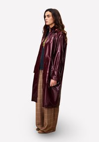 Bordeauxwaterdichte jas met een glanzende afwerking, lange mouwen en een capuchon; gecombineerd met gemusterde bruin geruite broek en beige schoenen.