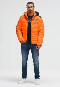 Bright orange puffer jacket with navy accents, quilted texture, front zipper, and printed details. Worn over a navy t-shirt and blue jeans.
