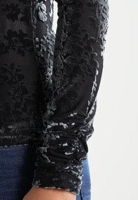 Svart sammet långärmad topp med blommönster, rynkade ärmar och en genomskinlig överlagring, matchad med blå denim.