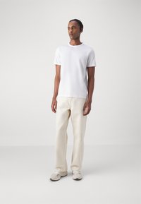 Weißes Baumwoll-T-Shirt, kurze Ärmel, Rundhalsausschnitt, kombiniert mit hellbeigen Hosen. Sneakers in Hellgrau mit weißen Akzenten. Schlichtes Design.