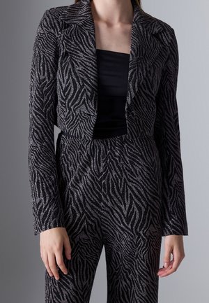 Femme portant un blazer noir et argenté à motif zèbre et un pantalon taille haute assorti sur un top noir, debout devant un fond gris.