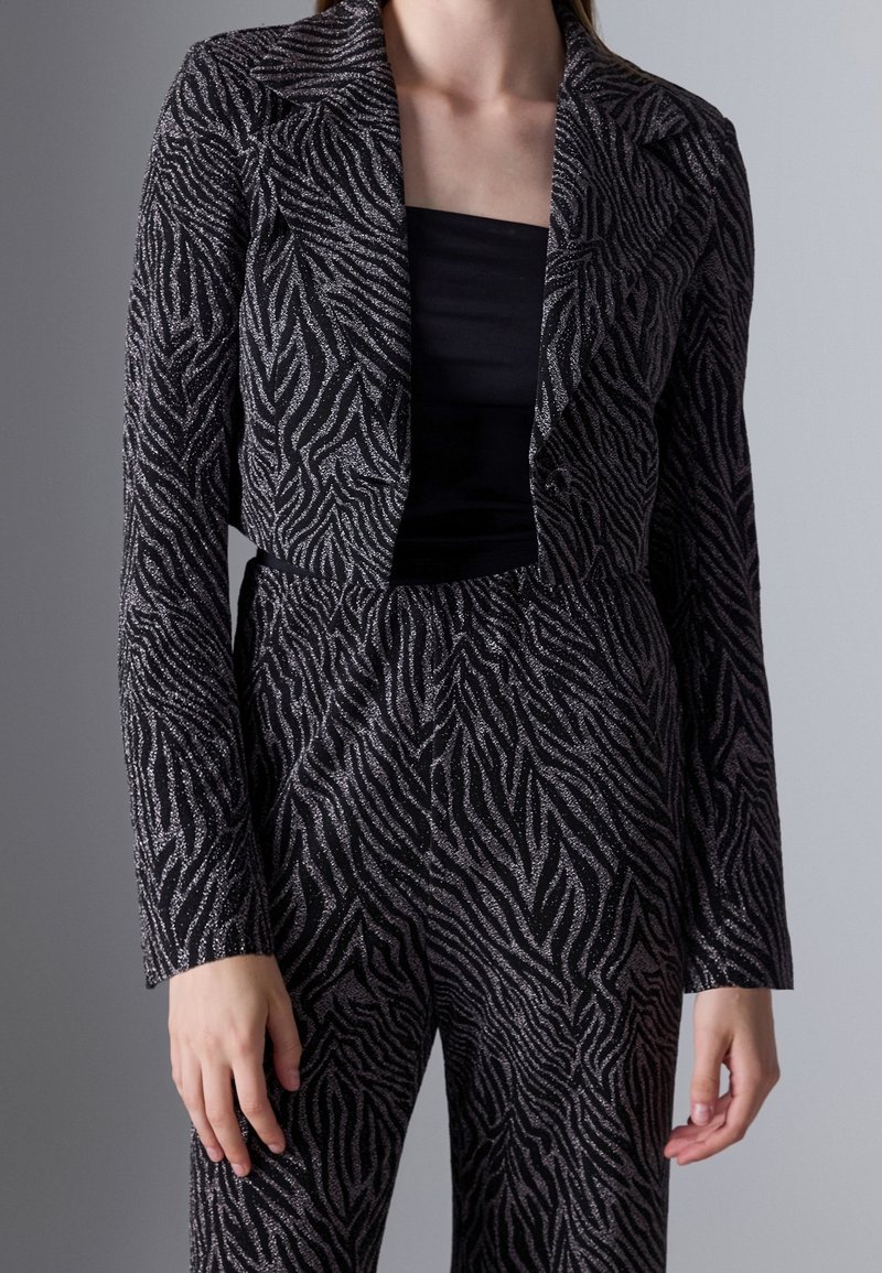 Femme portant un blazer noir et argenté à motif zèbre et un pantalon taille haute assorti sur un top noir, debout devant un fond gris.