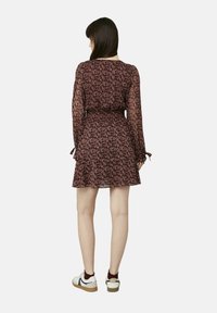 Robe à motif maroon avec de longues manches transparentes, taille froncée et ourlet en volants. Associée à des baskets blanches et des chaussettes bordeaux. Vue de dos.