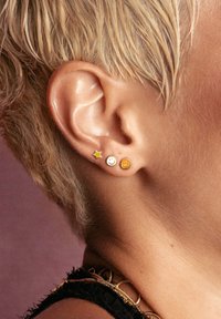 Pendientes en forma de estrella dorada y dos caras sonrientes en la oreja de un modelo; los pendientes presentan una superficie brillante con formas distintivas y sutiles variaciones de color.