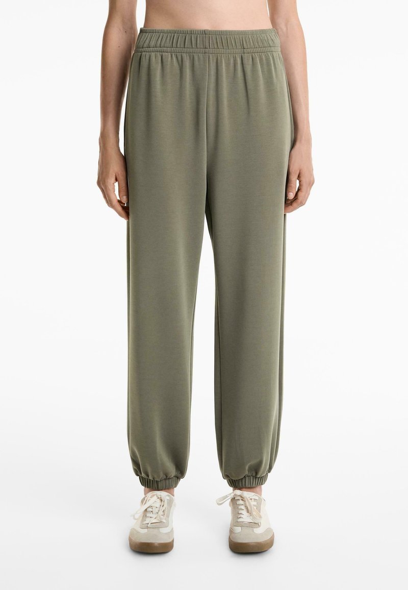 Lys olivengrønne sweatpants med elastisk talje og manchetter, lavet af blødt stof. Båret med beige sneakers med hvide detaljer.