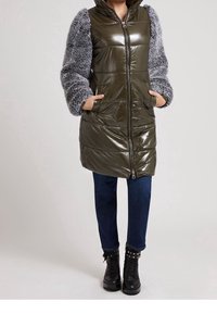 Donna che indossa un giubbotto puffer lucido di colore verde oliva con maniche grigie e morbide, jeans blu scuro e stivali neri con borchie e lacci, con le mani in tasca.