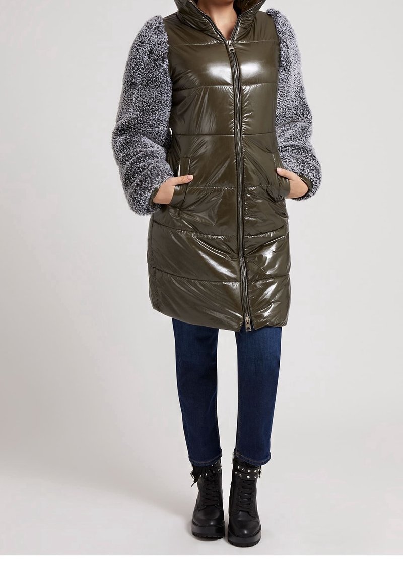 Donna che indossa un giubbotto puffer lucido di colore verde oliva con maniche grigie e morbide, jeans blu scuro e stivali neri con borchie e lacci, con le mani in tasca.