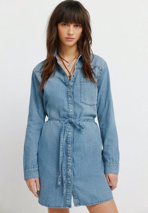 Kvinde iført en denimskjortekjole med bælte og lange ærmer samt lagdelte halskæder, stående foran en ensfarvet hvid baggrund.