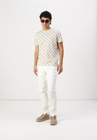 Chemise beige à manches courtes avec un motif géométrique, associée à un pantalon blanc et des chaussures marron clair. Le modèle porte des lunettes de soleil noires.
