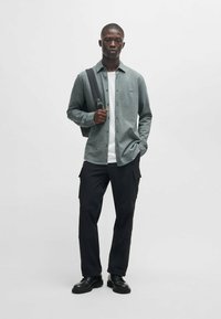 Grijze button-up shirt over een witte t-shirt, gecombineerd met zwarte cargo broek en zwarte veterschoenen. Model houdt een zwarte schoudertas vast.