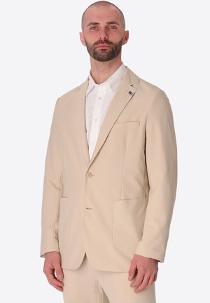 Uomo con barba che indossa una giacca da abito beige e pantaloni abbinati sopra una camicia bianca abbottonata, in piedi su uno sfondo semplice.