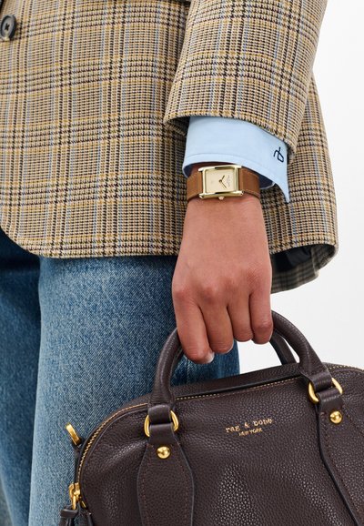 Hnědá kožená kabelka se zlatými doplňky držená v ruce, oblečená v kostkovaném saku a světle modré košili, doplněná zlatými čtvercovými hodinkami.