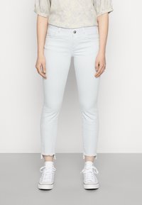 Světle modré, úzké džíny s roztrženým lemem. Vyrobené z denimu, obsahují pět kapes a zapínání na přední knoflík. Nohavice jsou obuty s bílými teniskami.